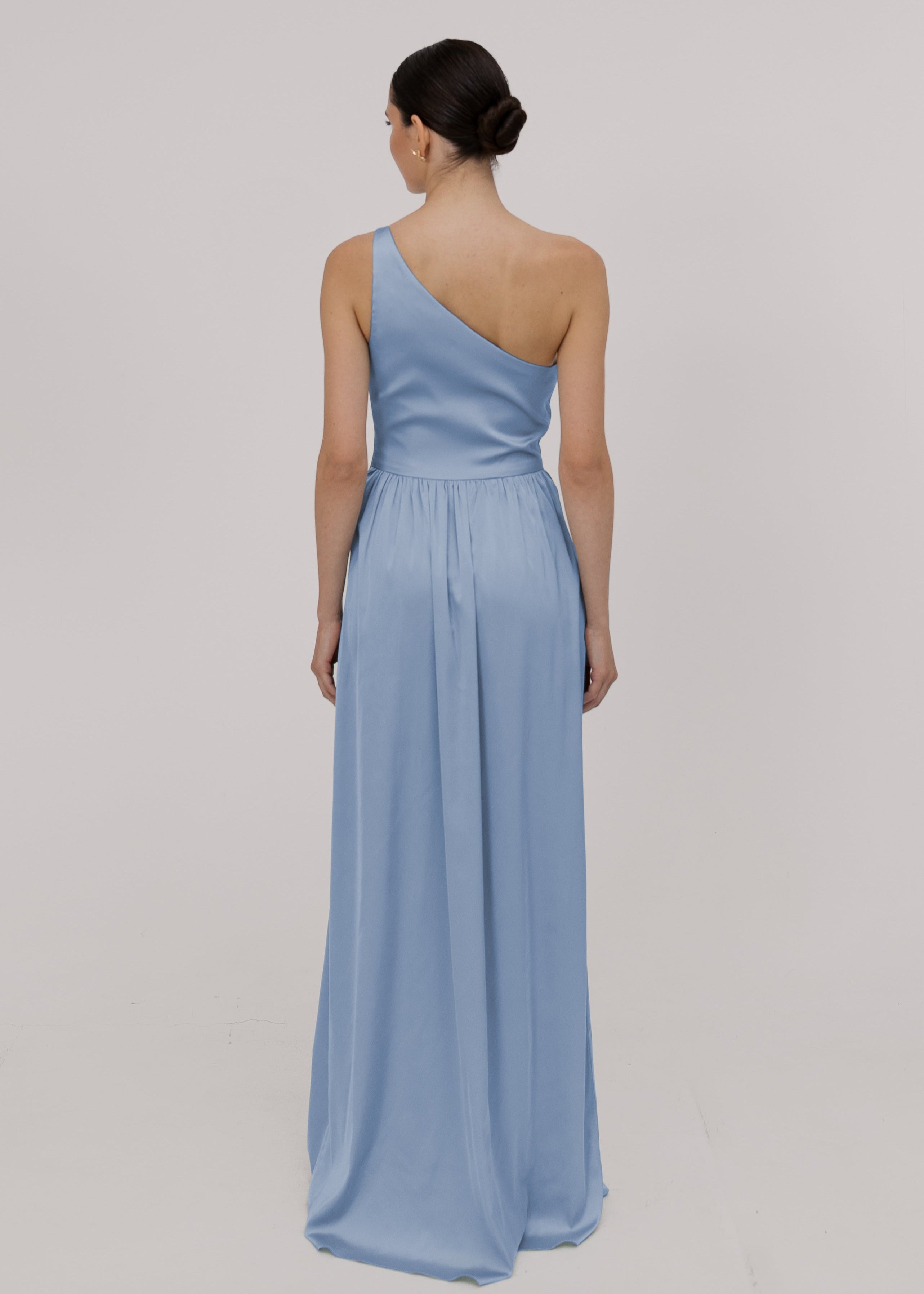 Model wearing a floor length dusty blue satin bridesmaid dress with one shoulder and slit in skirt by TH&TH Bridesmaids.
