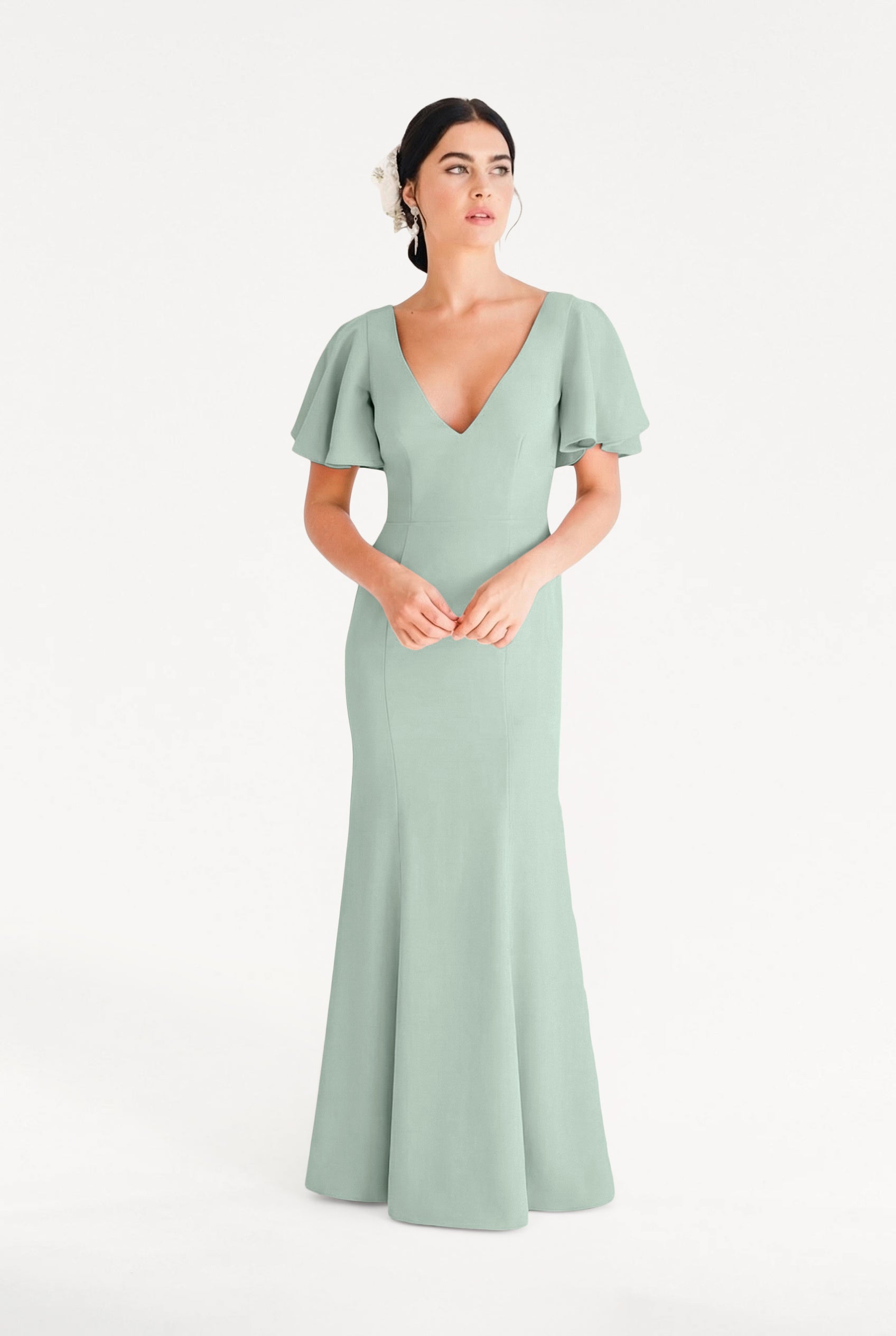 A woman stands against a white background wearing the TH&TH Celeste Moss Crepe Bridesmaid Dress in Sage Green, featuring flutter sleeves and a deep V-neckline. Her dark hair is styled back with a white flower accessory.