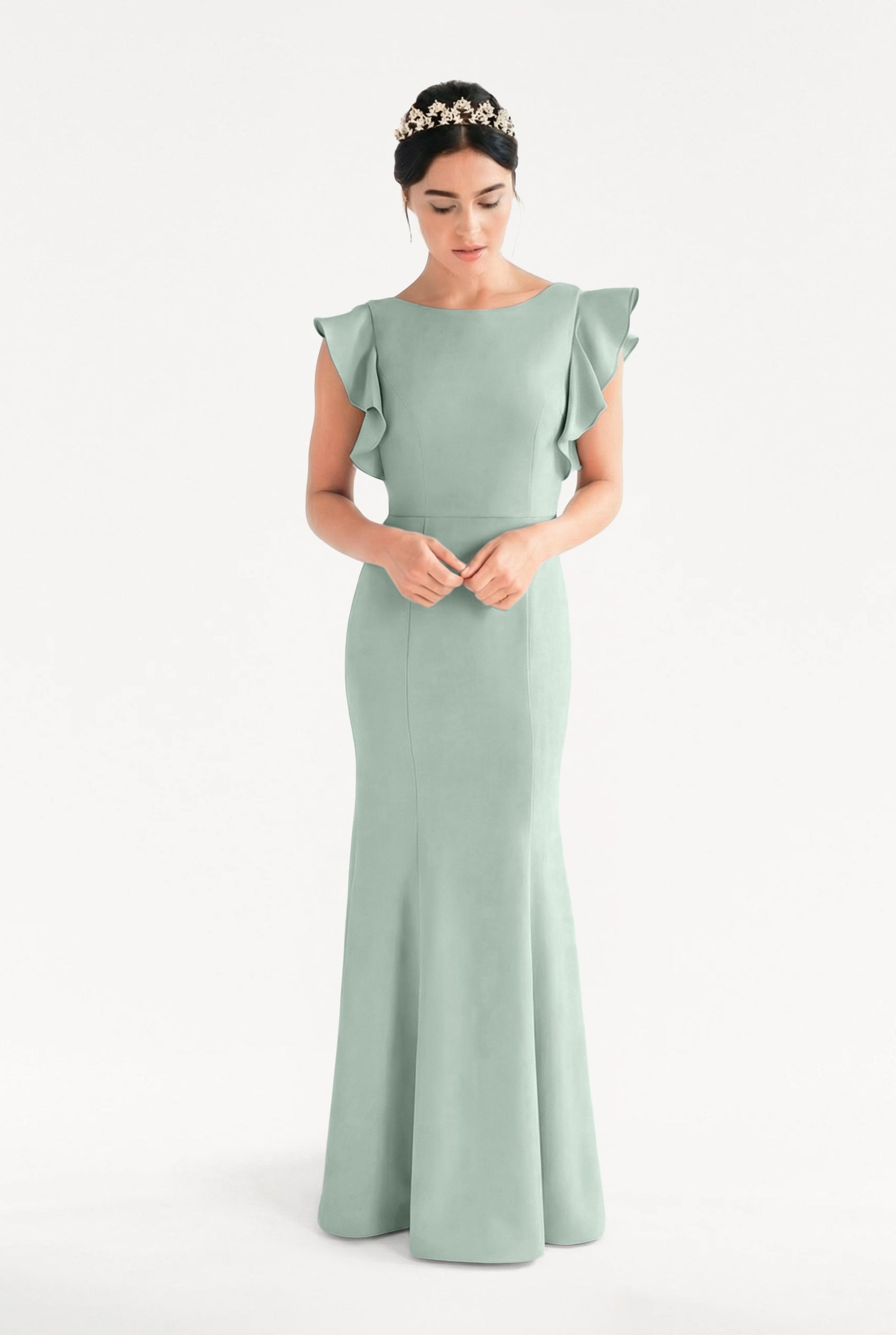 A woman stands against a white background wearing the TH&TH Cecelia Moss Crepe Bridesmaid Dress in Sage Green, a floor-length gown with ruffled cap sleeves. She clasps her hands and wears a floral headband while looking down.