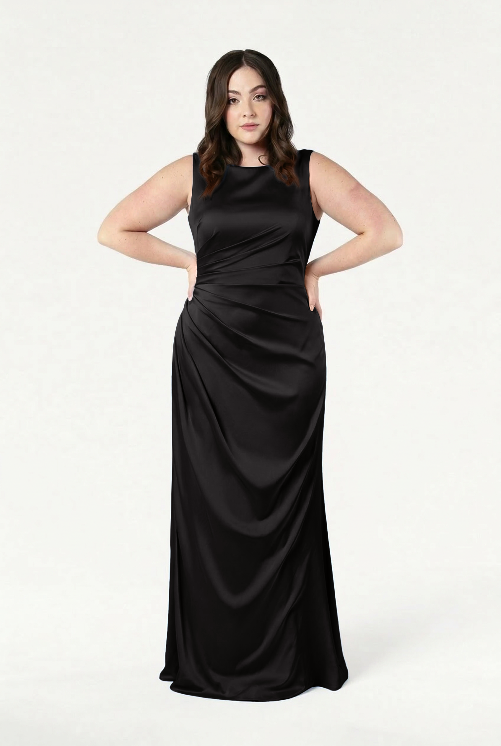 A woman with long brown hair stands confidently against a white background, wearing the TH&TH Nala Bridesmaid Dress in Black Satin—an inclusive, floor-length gown featuring ruched details and Grecian-inspired pleating.