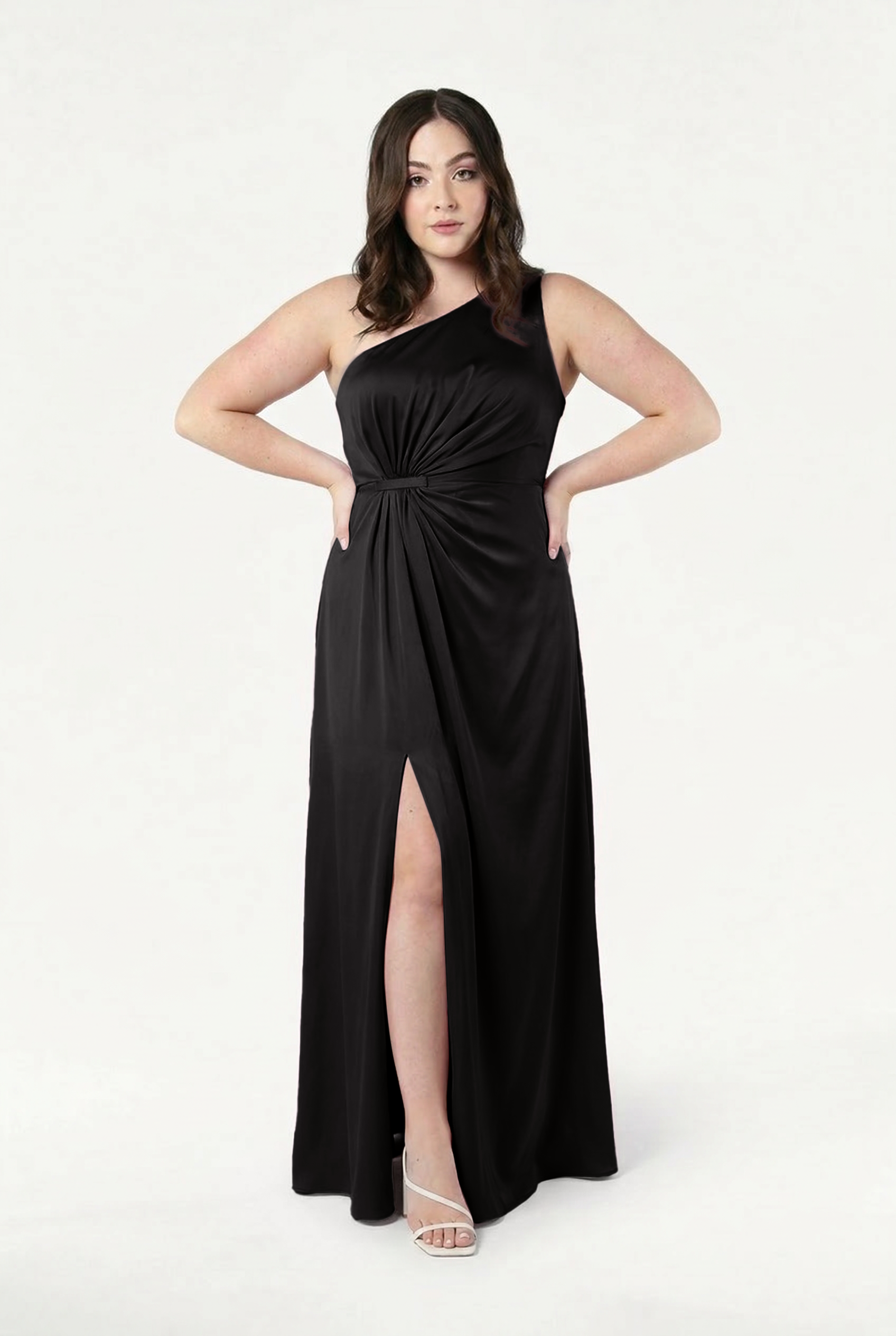 A woman models the TH&TH Greta Bridesmaid Dress in Black Satin with Grecian side draping, posing confidently with one hand on her hip against a white background, paired with white heeled sandals for an elegant look.
