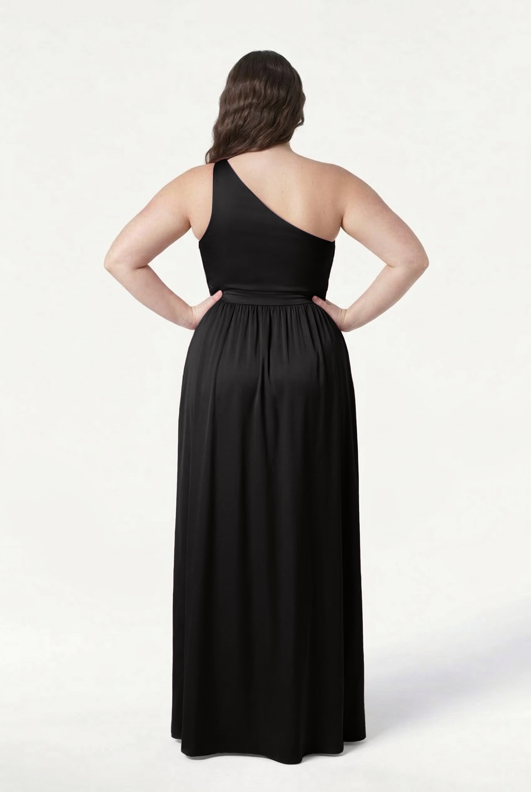 A woman stands with her back to the camera, hands on hips, modeling the TH&TH Greta Bridesmaid Dress in Black Satin, featuring a one-shoulder design, fitted waist, and flowing skirt against a plain white background.