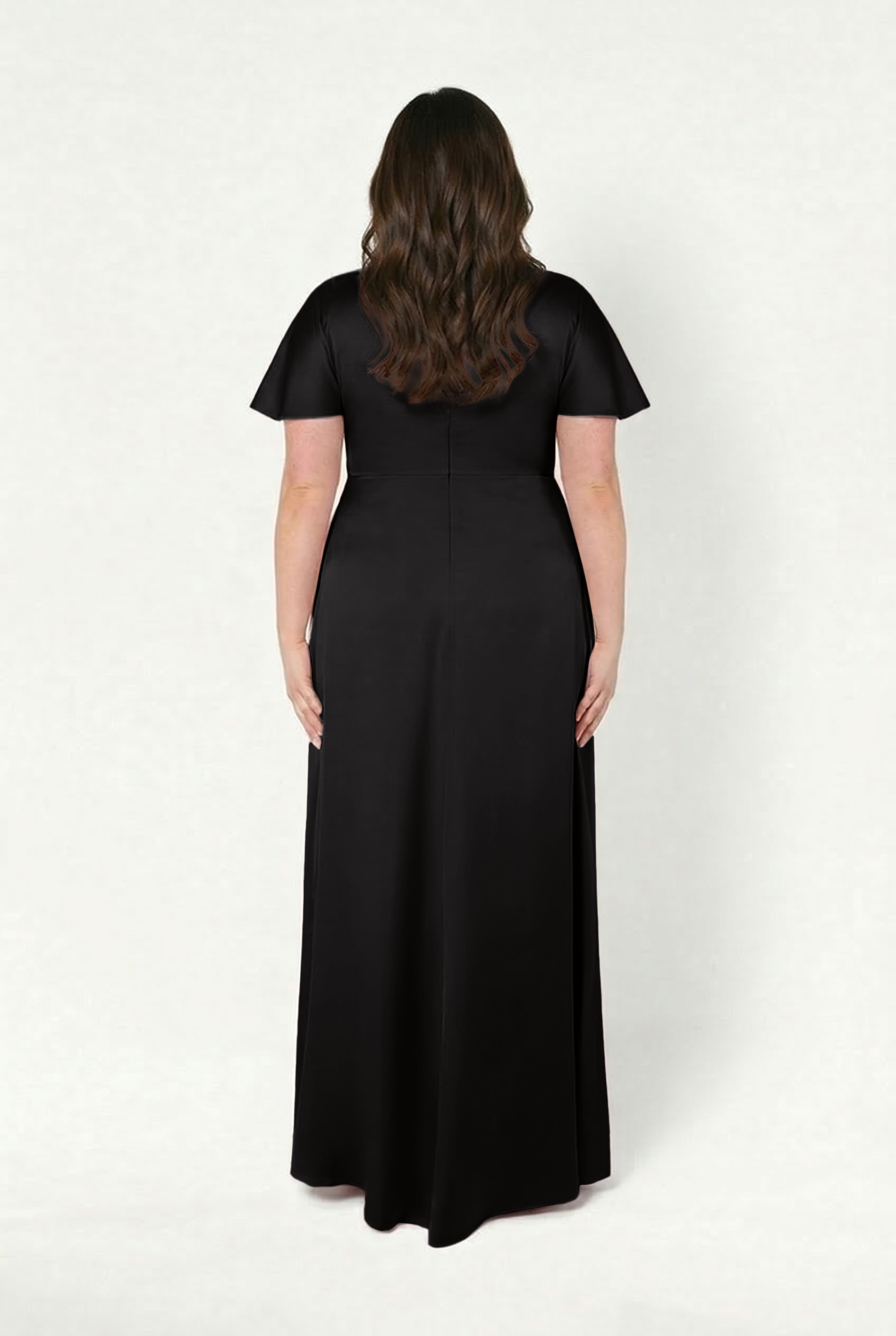 A woman with long, wavy brown hair faces away, modeling the TH&TH Camilla Bridesmaid Dress in Black Satin—a floor-length, short-sleeve gown—against a plain white background.