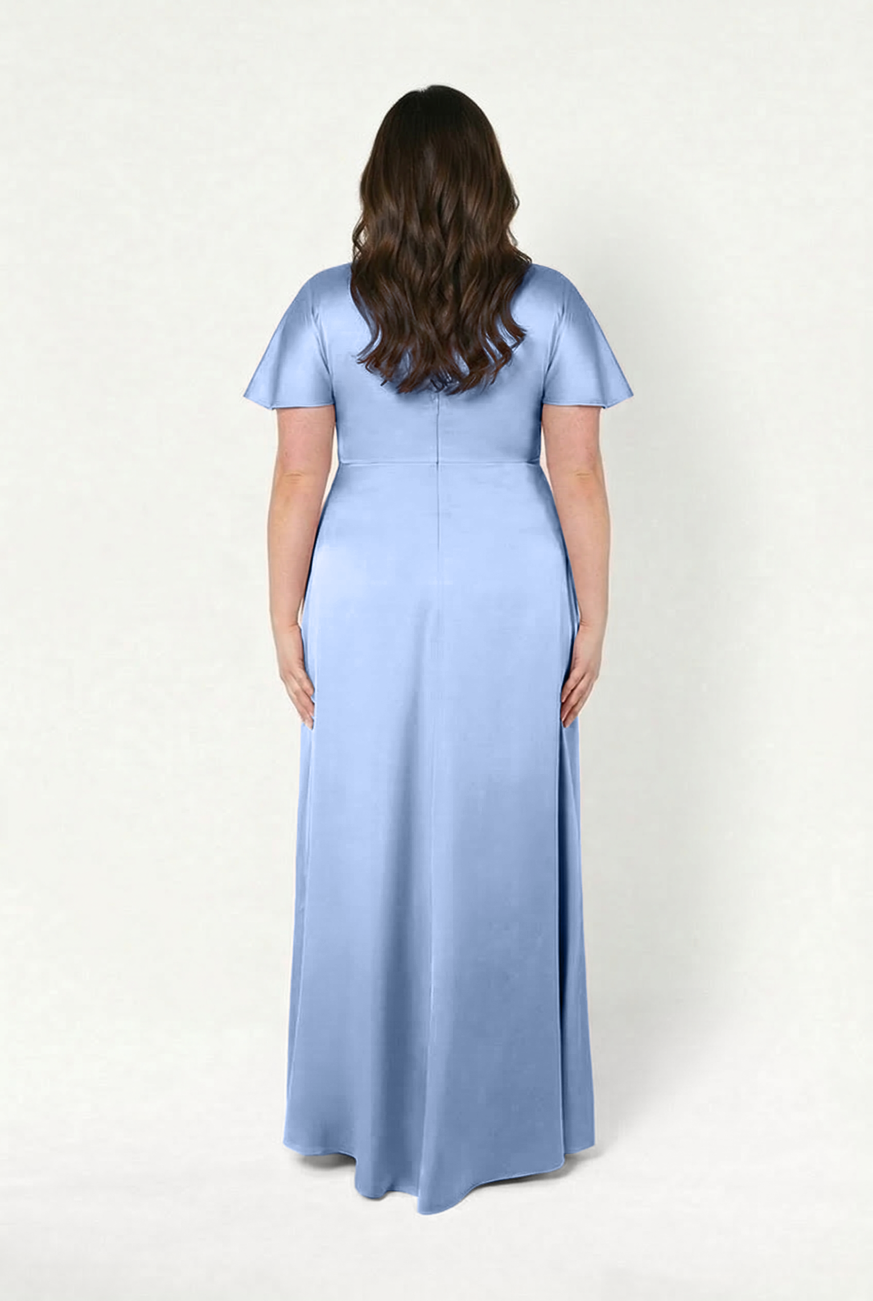 A woman with long, wavy brown hair stands with her back to the camera, wearing the TH&TH Camilla Bridesmaid Dress in Dusty Blue Satin. The floor-length dress has short sleeves and is set against a plain, light background.