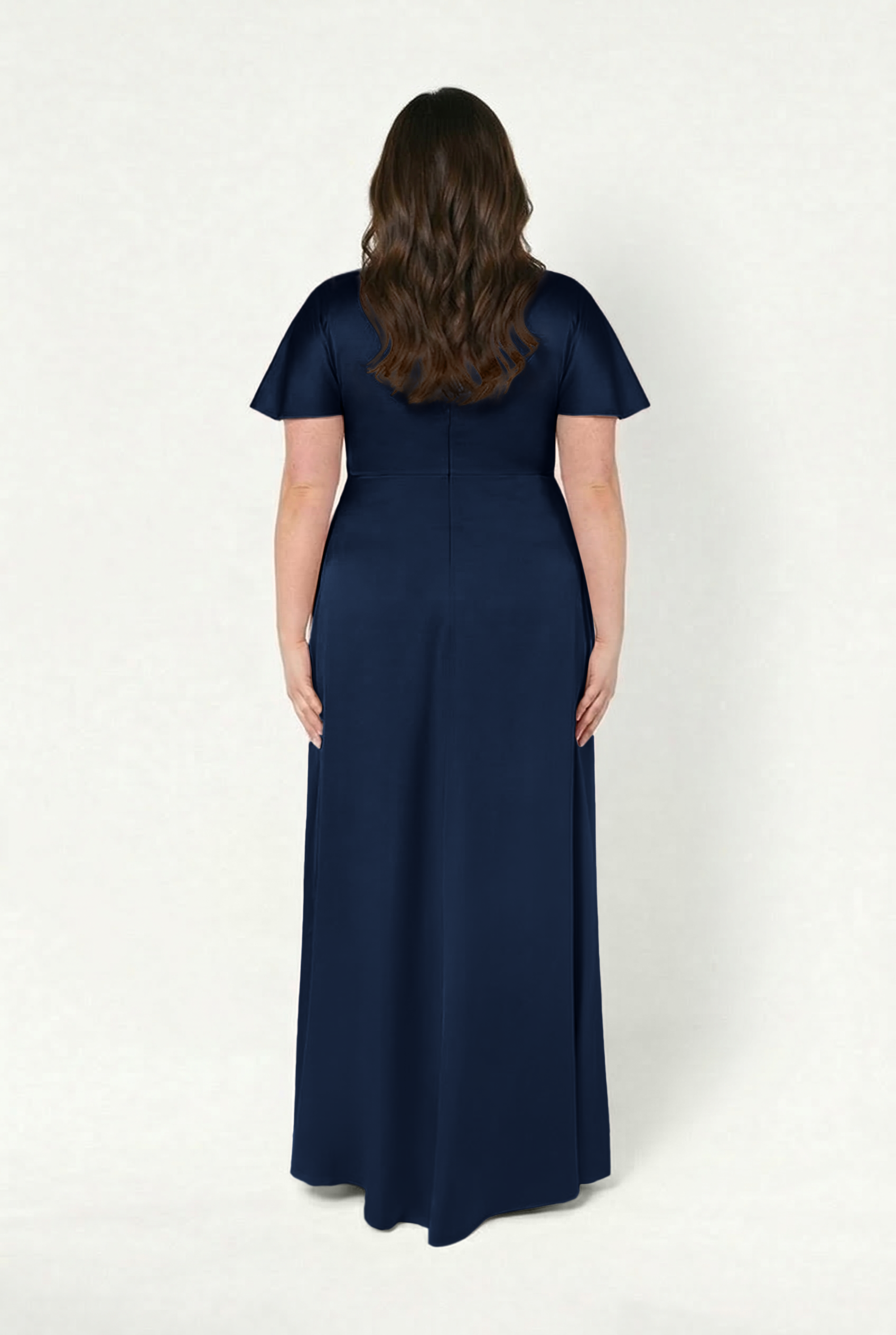A woman with long wavy brown hair stands with her back to the camera, wearing the TH&TH Camilla Bridesmaid Dress in navy satin, which features a floor-length design and a twist-knot bodice against a plain background.